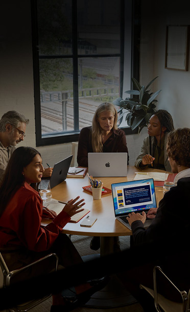Vijf personen zitten rondom een vergadertafel en werken samen op hun MacBook, met iPhones met het scherm omlaag op de tafel