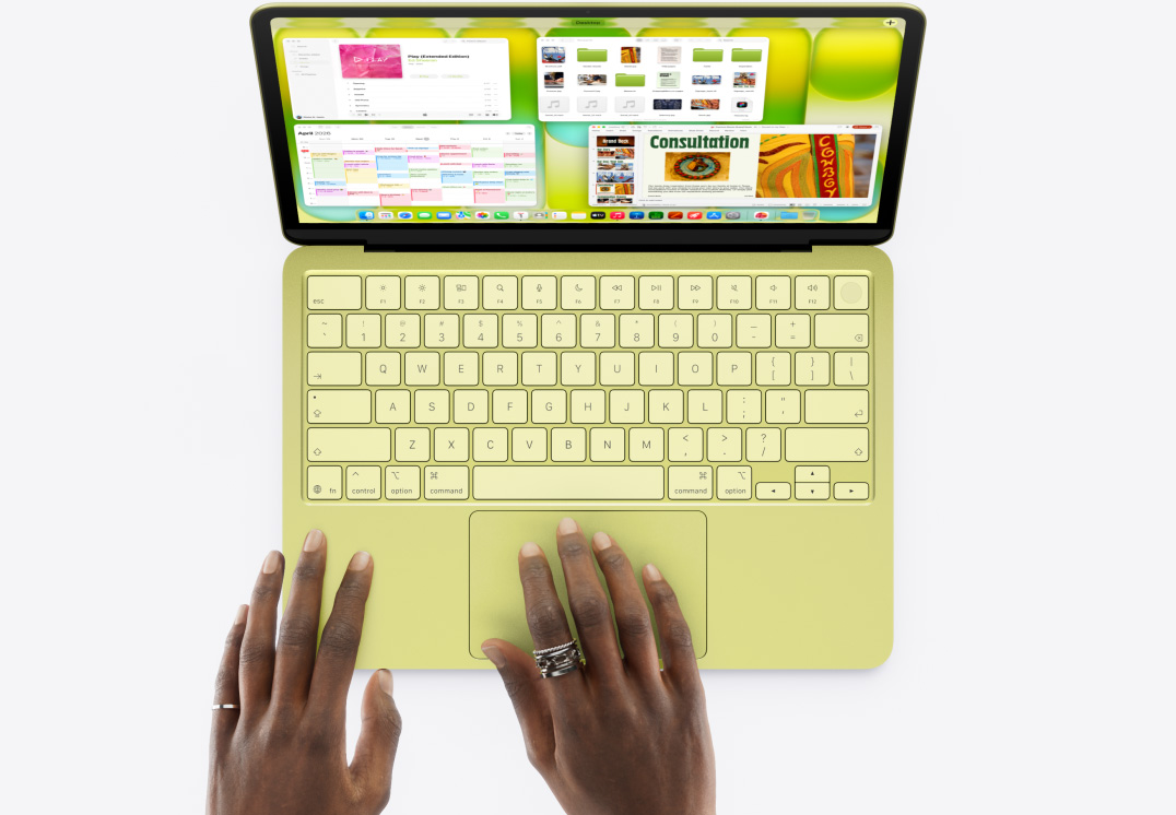 MacBook Neo, citrusová farba. Na trackpade je ruka, otvorených je viacero okien vrátane apiek Hudba, Kalendár a Microsoft PowerPoint.