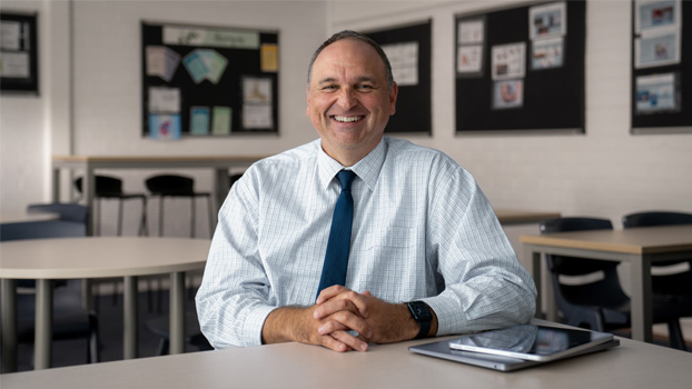 Le proviseur Mark Watkins souriant, assis à une table de classe, avec un MacBook fermé et un iPad à côté de lui.