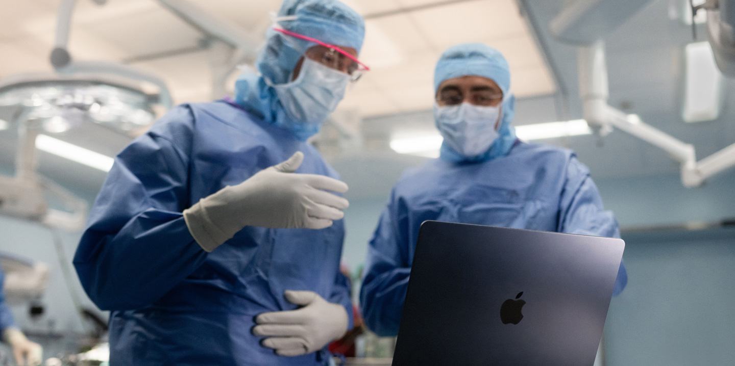 Hai bác sĩ phẫu thuật trong phòng mổ nhìn vào MacBook, một người ra hiệu bằng tay trong khi người kia quan sát