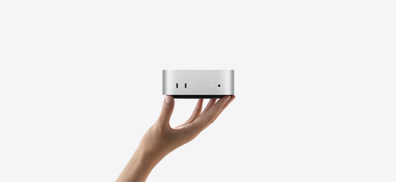 Front view of Mac mini balanced on the fingertips of an up-stretched hand, front shows two Thunderbolt ports, status indicator light and headphone jack, tapered black base at bottom, flat top, rounded sides, straight edges, silver color